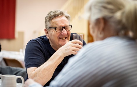 A picture of an older man laughing. He is wearing a t-shirt and has glasses. His eyes are closed and creased up from laughing and he has a big grin, showing his teeth. The person sitting opposite him must have said something that was very funny!