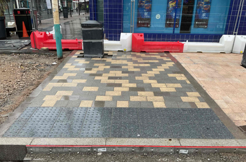 A photo of the paving slabs on Eastbank Street. There are tactile paving slabs for the crossing, and the smooth tiles are grey, yellow and black. To the right, the paving tiles not at the crossing are pink.