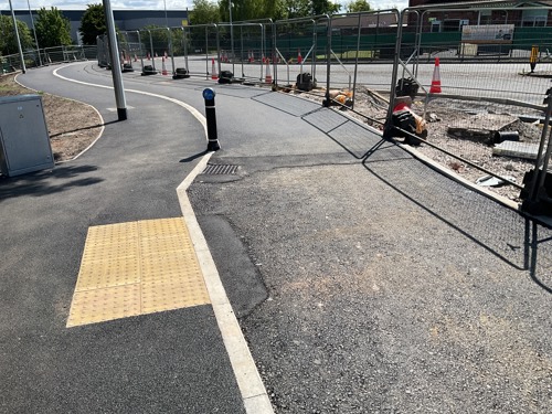 A photo of a new section of crossing, with a bollard, textured paving slaps marking the crossing. There is fencing to the right, showing works are continuing.