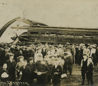 Sepia photo of crowd stood in front of wrecked train carriage